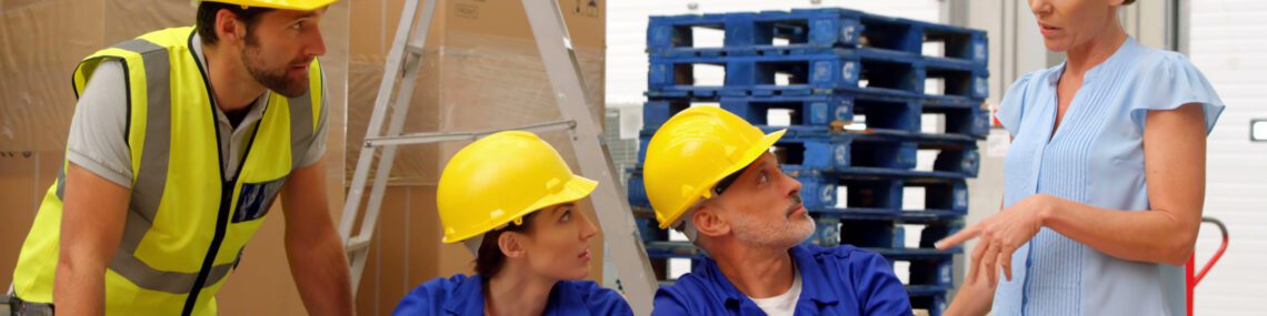 Group of warehouse employees holding team meeting. Female manager providing instructions to diverse team members gathered around desk with laptop. Ideal for illustrating concepts of teamwork, leadership, logistics, and organizational communication in industrial settings. Useful for training materials, business presentations, and articles on effective workplace interactions.