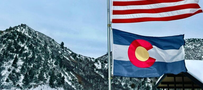 Colorado flag beneath American flag with mountains in background