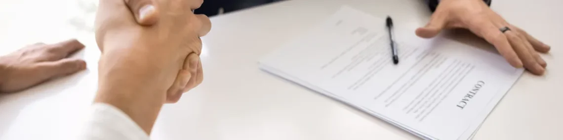 Buyer and seller shaking hands across a table with a signed business purchase contract and pen