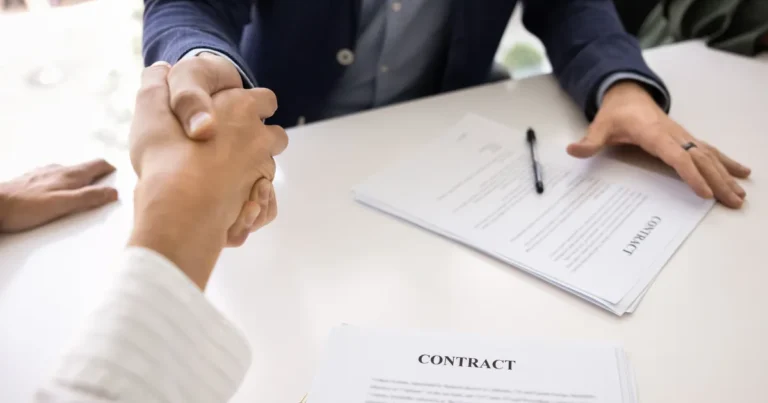 Buyer and seller shaking hands across a table with a signed business purchase contract and pen