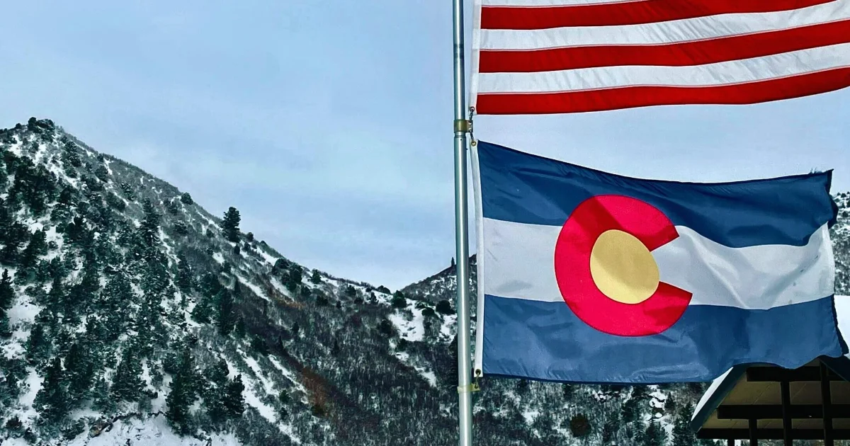 Colorado state flag flying beneath the American flag with snowy mountains behind