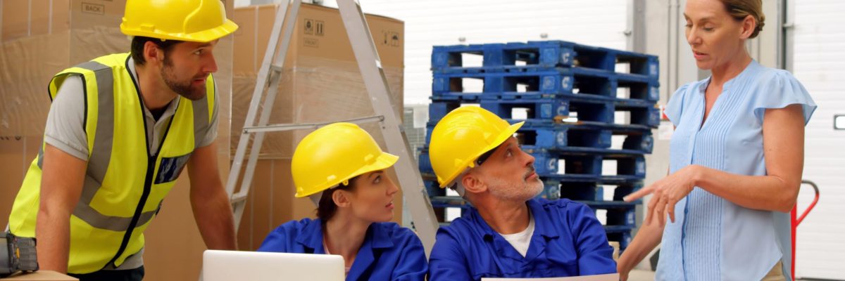 Group of warehouse employees holding team meeting. Female manager providing instructions to diverse team members gathered around desk with laptop. Ideal for illustrating concepts of teamwork, leadership, logistics, and organizational communication in industrial settings. Useful for training materials, business presentations, and articles on effective workplace interactions.