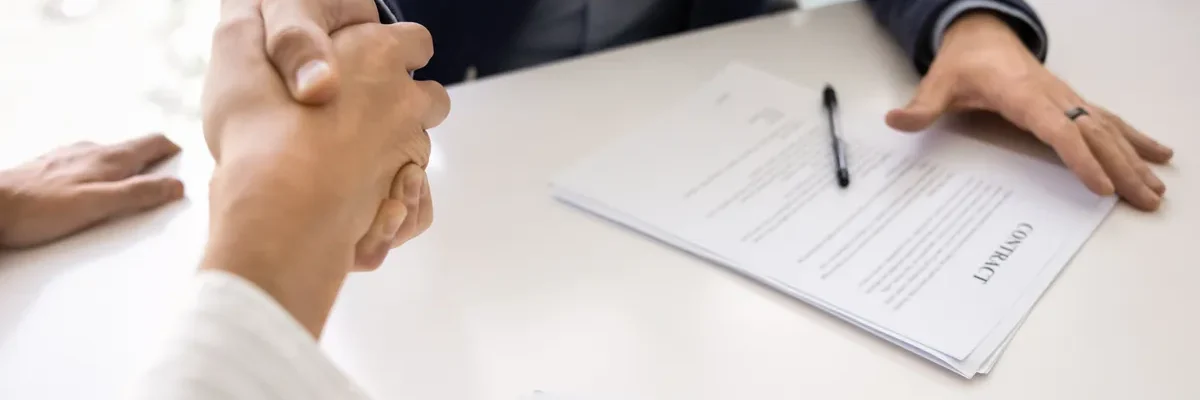Buyer and seller shaking hands across a table with a signed business purchase contract and pen